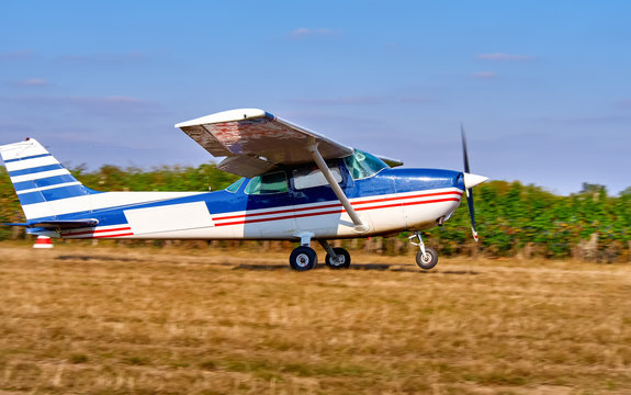 A light aircraft takes off in a field near a vineyard. Sunny summer day, people watch and plant.