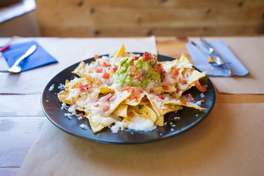 black plate with nachos and guacamole, chopped tomato and cheese, on wooden table with paper placemats at restaurant
