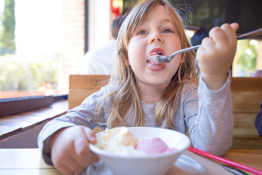 Portrait Of Three Years Old Blonde Child Eating With Spoon Two Scoops Of Strawberry And Vanilla Ice Cream, In White Bowl, Sitting At The Restaurant Table
