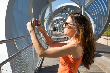 adult woman with orange shirt in summer, taking a photo or video recording with a smartphone, at modern urban footbridge, in Madrid city, Spain, Europe
