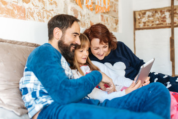 Couple relaxed at home in bed with their little daughter on the mobile phone and tablet.