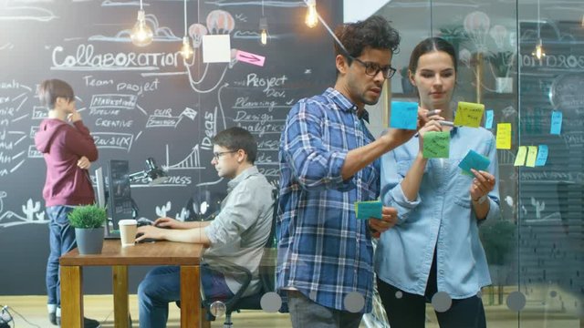 Creative Male and Female Developers Use Glass Wall for Outlining New Project with Sticky Papers. In the Background Talented Young People Working with Blackboard and on Personal Computer. 