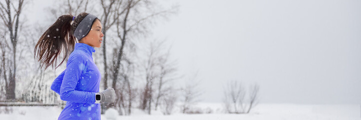 Winter running smartwatch woman in cold snow weather jogging outside wearing windproof clothes with gloves, headband, winter tights and wind jacket in white snowfall background.