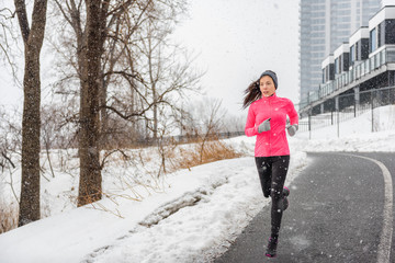 Winter running Asian girl wearing cold weather clothing for outside exercise in snow storm snowfall during winter training outdoors in city street. Fitness woman exercising.