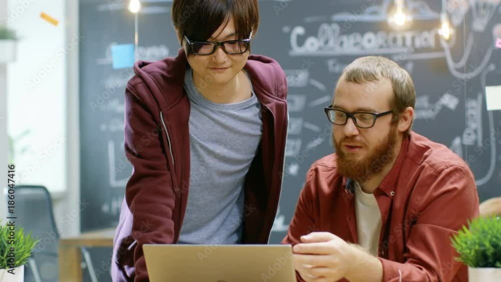 Poster engineer talks with developer about ongoing project while he works on a laptop. in the background st