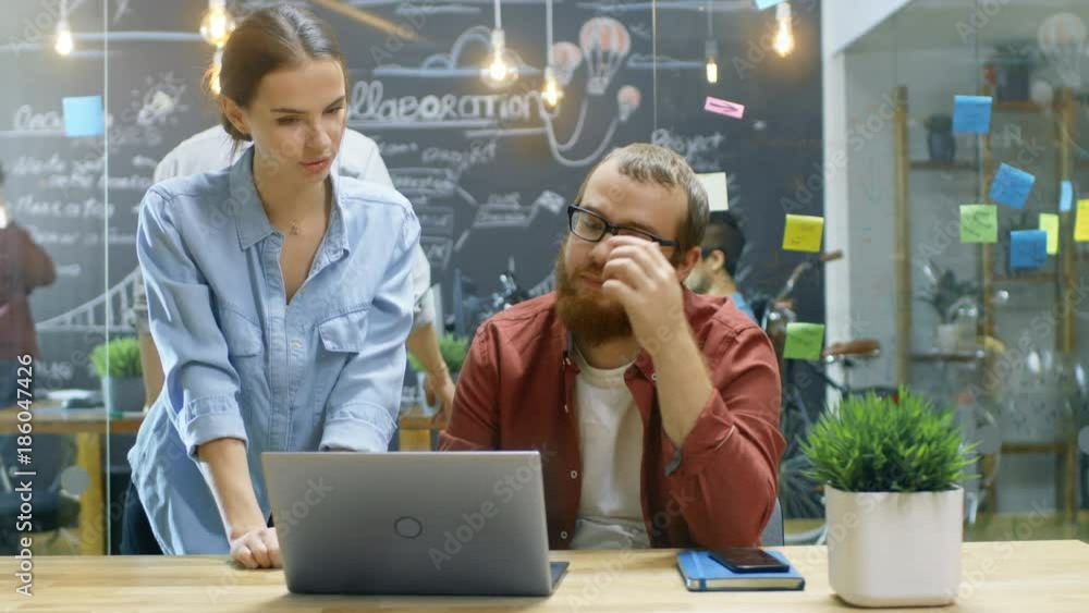 Poster female designers talks male developer about ongoing project while he works on a laptop. in the backg