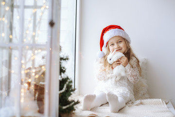 cute little girl in a white dress with a white little rabbit in a santa claus hat