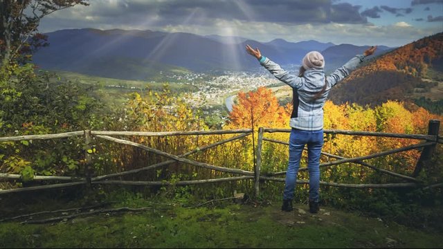 Young woman standing on the edge of the cliff and raising her hands up against mountain range and bright colorful autumn forest shining in sun rays. Success and travel concept. Toning filter. Full HD