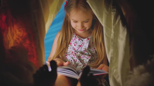 Cute little girl is reading a book in tent. Happy girl plays at home. Funny lovely kid having fun in children room.