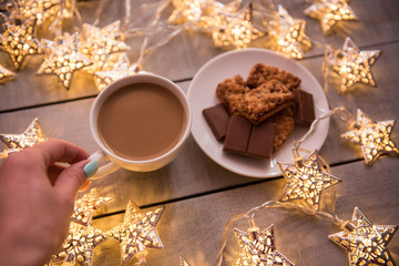 Fototapeta na wymiar Christmas and new year holiday celebration concept background. Cup of coffee, homemade chocolate cookie and peanut biscuit, lighted candles, xmas tree decoration on wooden table.