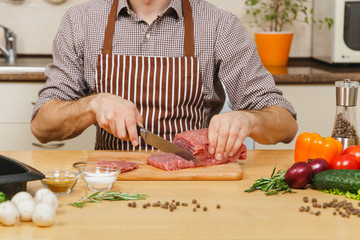 Close up caucasian young man in apron sitting at table with vegetables, cooking at home preparing meat stake from pork, beef or lamb, in light kitchen with wooden surface, full of fancy kitchenware.