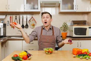 Fun caucasian young man in apron, brown shirt sitting at table, choosing between onion and tomato,...