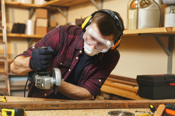Caucasian young man in plaid shirt, black T-shirt, noise insulated headphones, protective mask working in carpentry workshop at wooden table place with different tools, sawing iron with power saw.