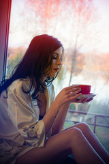 Naklejka premium Young beautiful dreaming brunette woman drinking cup of coffee wearing lingerie sitting home by the window. Blurred yard tree background, concept of lady's dreams and wishes 