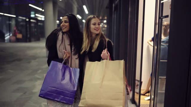 Two cute young adolescent women, heavy loaded with christmas gifts,  shopping, while walking along store windows and choosing and admiring the items. 