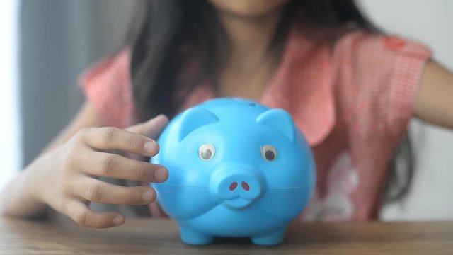 Asian little girl in Thai kindergarten putting money coin into clear piggy bank on wooden table 