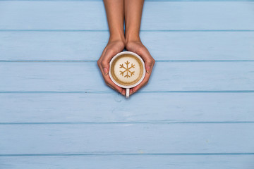 Woman's hands hold cap of coffee with snowflake ornament on a blue wooden table, view above. 