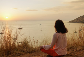 Young girl sitting in yoga lotus meditation position in front to seaside on the rocks an watching...