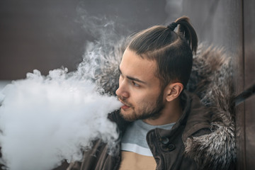 Vape man. Portrait of a handsome young white guy with modern haircut vaping an electronic cigarette opposite the futuristic urban background. Lifestyle.