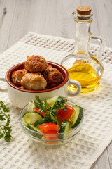 Fried meat cutlets in ceramic soup bowl, bottle with sunflower oil and salad of fresh cucumbers and tomatoes in glass bowl on white kitchen towel