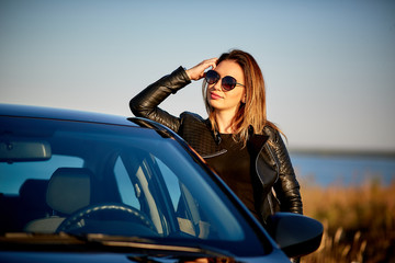 Fototapeta na wymiar beautiful young woman in a leather jacket and sunglasses stands near the car