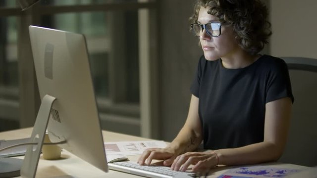 Young businesswoman in eyeglasses typing on computer keyboard and reading documents at office workplace