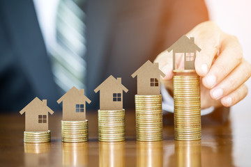 Businessman hand holding money coin with wooden toy house putting on money coins stack arranged as a graph on wooden table, concept of money growth and saving money
