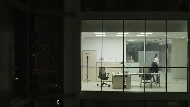 View from outside of modern glass building of businessman entering office, turning on light and start working on computer at night