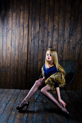 Elegante blonde girl wear on fur coat sitting on chair at studio against wooden background.
