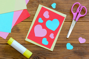 Nice Valentines day card with paper hearts, scissors, glue stick, colored paper sheets on a wooden...
