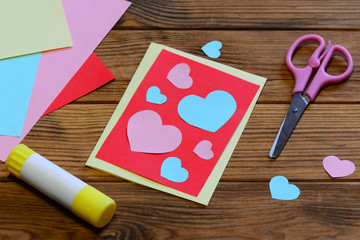 Valentines day greeting card with paper hearts, scissors, glue stick, paper sheets on a wooden...