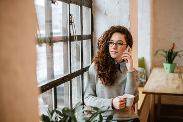 Pretty woman talking on phone near window at home and holding cup.