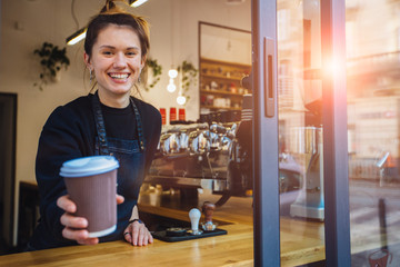 Smiling friendly barista holding disposable cup at coffee shop. Coffee business concept. - obrazy, fototapety, plakaty