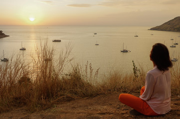 Young girl sitting in yoga lotus meditation position in front to seaside on the rocks an watching the sun goes down in a golden hour. sunset over sea.