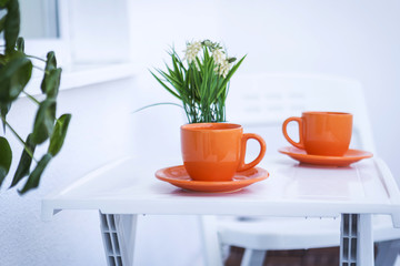 orange cups with hot coffee or tea served on a white table. an arranged table with glasses and fresh green flowers. pleasant breakfast on the veranda.