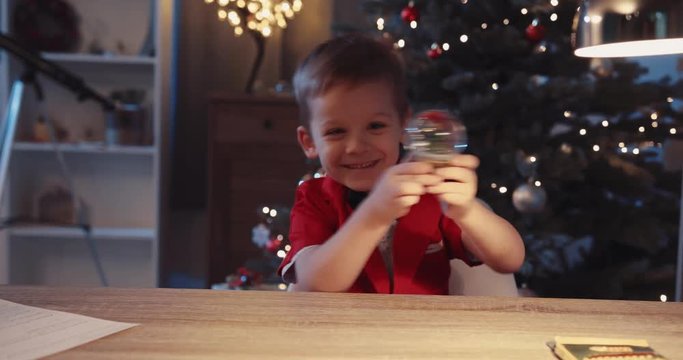 Smiling little boy sit on the table smile hold play small toy minature near Christmas tree in home figurine globe holidays joy kids lights child holiday happy portrait close up winter slow motion