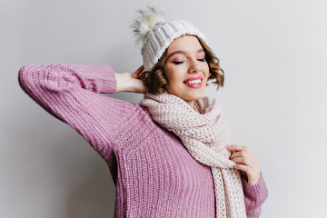 Short-haired woman with pink makeup posing on light background in knitted hat. Studio shot of dreamy curly female model in purple sweater and white scarf.