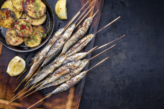 Traditional Spanish barbecue sardines on a wooden skewer with fries as top view on a cutting board