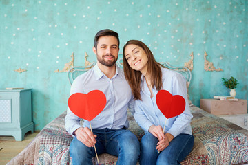 A couple with hearts in their hands sitting on the bed laughing indoors. Valentine's Day.