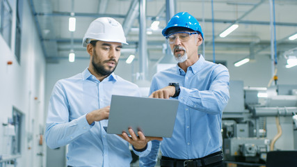 Head of the Project Holds Laptop and  Discusses Product Details with Chief Engineer while They Walk Through Modern Factory.