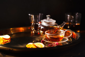 Small glass teapot and glasses with hot black tea, dried rose petals, pocket magnifier on golden chain, squeezed orange slice on golden tray. Evening light. Dark background