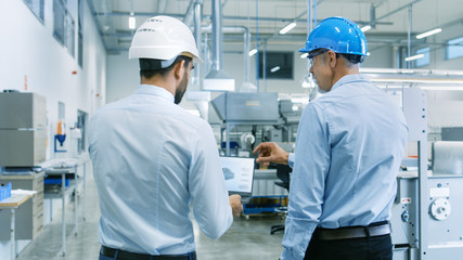 Back View of the Head of the Project Holds Laptop and Discussing Product Details with Chief Engineer while They Walk Through Modern Factory.
