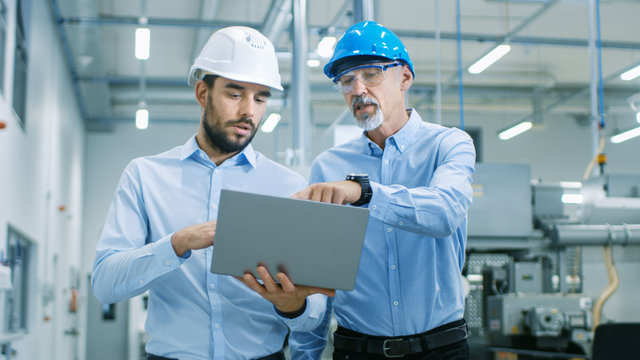 Head of the Project Holds Laptop and  Discusses Product Details with Chief Engineer while They Walk Through Modern Factory.