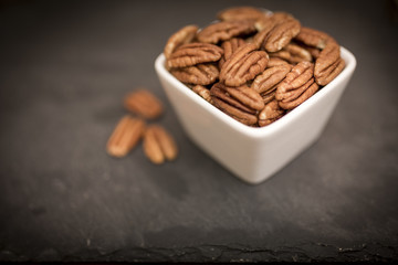 A white bowl of healthy and nutritious pecan nuts shot from above on a black slate background
