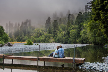 Couple Sits on Footbridge Over Calm Mountain River