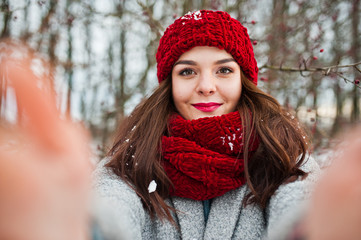 Portrait of gentle girl in gray coat , red hat and scarf near the branches of a snow-covered tree, holding camera for selfie.