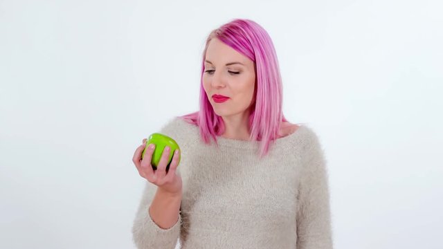 A young woman with pink hair is biting into a delicious green apple.
