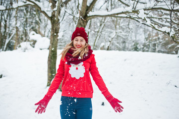 Blonde girl in red scarf, hat and santas sweater posing at park on winter day.