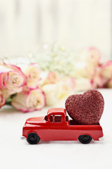 Obraz na płótnie Canvas 1950's antique vintage red truck hauling a glittery red heart past a roll of beautiful long stem roses. Extreme shallow depth of field with selective focus on vehicle.