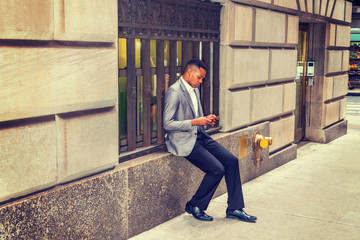 African American man reading, texting on cell phone on street, traveling, working in New York, wearing gray blazer, black pants, leather shoes, sitting on vintage window frame. Filtered look..